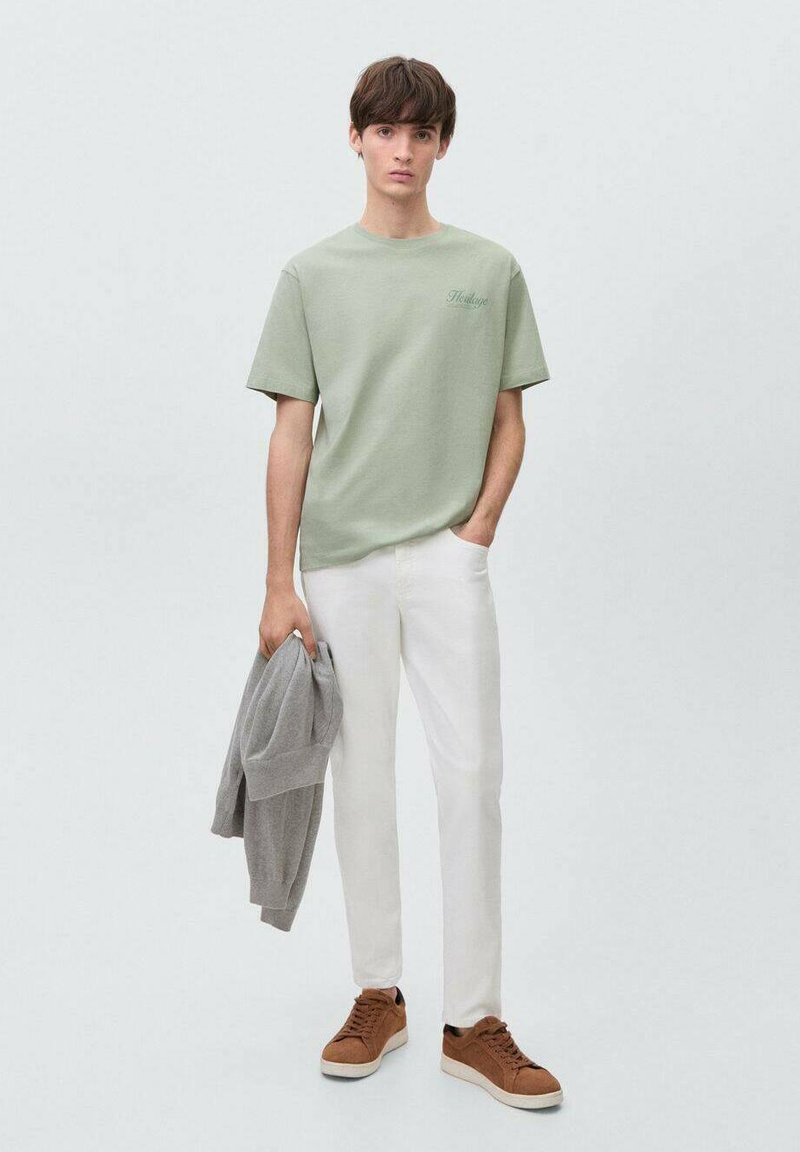 Camiseta de algodón verde claro con mangas cortas, zapatillas color beige y pantalones blancos. Sosteniendo un suéter gris, de pie sobre un fondo neutro.