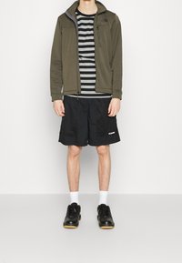 Olivově zelená fleecová bunda přes černé a šedé pruhované tričko, doplněná černými šortkami a černými teniskami. Viditelné bílé ponožky.