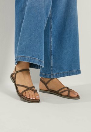 Piedi con sandali bassi marroni con cinturini abbinati a jeans blu a gamba larga su uno sfondo chiaro e semplice.