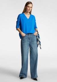 Blauwe v-hals trui, losse pasvorm, gecombineerd met lichtblauwe wijde jeans. Opvallende gestreepte sjaal gestrikt om de taille. Zwarte puntige schoenen.