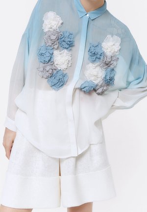 Blouse transparente bleue et blanche avec des applications florales 3D en bleu, blanc et gris, associée à un short large blanc texturé.