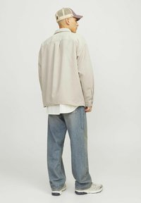 Beige corduroy overshirt with a collar, worn over a white striped shirt, paired with loose blue jeans and a beige cap.