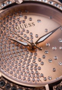 Rožnato zlato ura z teksturirano številčnico, ki vsebuje vzorec bleščečih kristalov, tanke kazalce in logotip "Guess".