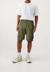 Olivgrön cargoshorts med sidorfickor, kombinerat med en löst sittande vit T-shirt och grå träningsskor med gula accenter.