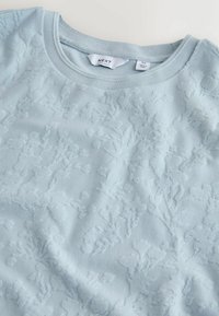 Pull bleu clair de taille moyenne avec un motif floral en relief subtil et un col rond, montrant les étiquettes de marque et de taille à l'intérieur du col.