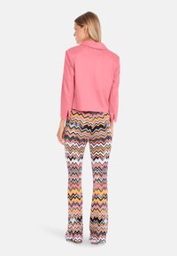 Veste courte rose avec un col, accompagnée de pantalons évasés à motif zigzag multicolore comprenant du noir, du blanc, du jaune et du rose.
