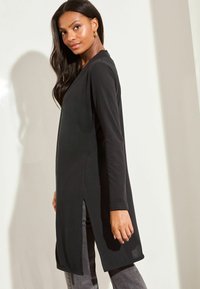 Fekete, hosszú ujjú tunika laza szabással, oldalán sliccel és puha anyagból. Szürke farmerral párosítva, kiemelve a laza megjelenést.