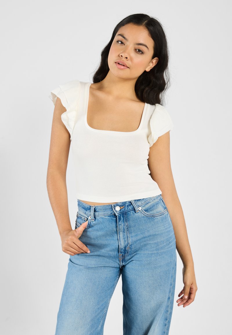 Hvit rippete crop topp med firkantet utringning, med korte, volangermede ermer. Sammen med høytlivte lyseblå jeans.