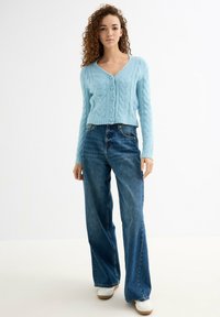 Cárdigan de punto azul claro con patrón de trenzas, cuello en V y botones frontales, combinado con pantalones vaqueros de pierna ancha azules y zapatillas blancas.