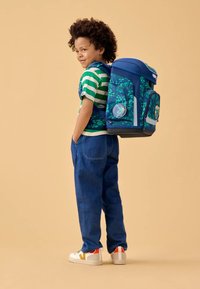 Blauer Rucksack mit Blättermuster, gepolsterten Trägern und Seitentaschen. Kind trägt ein grün gestreiftes Hemd und blaue Jeans, dazu weiße Sneaker.
