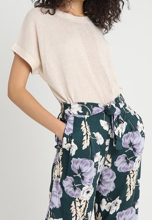 Mujer con un top de punto de manga corta color beige y pantalones de talle alto verde oscuro con un gran estampado floral en púrpura y blanco.
