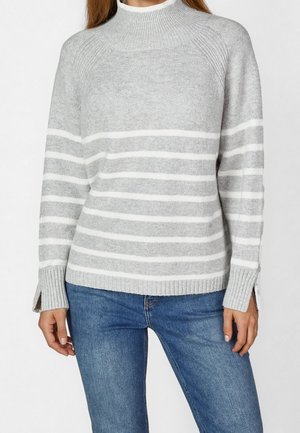 Femme portant un pull en tricot gris à rayures blanches horizontales et un jean bleu, debout avec les bras détendus sur les côtés.