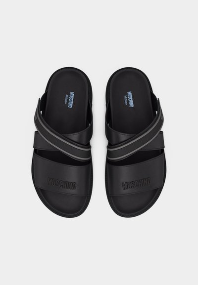 Sandales slides Noires Moschino Milano avec des brides croisées et un logo embossé sur les brides avant, vues de dessus sur un fond blanc.