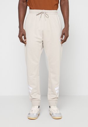 Homme portant un pantalon de jogging beige avec des bandes blanches près des chevilles et des baskets blanches, debout devant un fond clair uni.