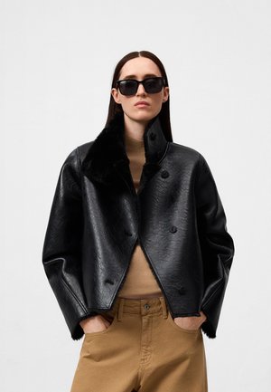 Zwartleren jasje met een nep bies, met een dubbel-breasted ontwerp en knoopsluitingen. Draagt over een beige top en tan broek.