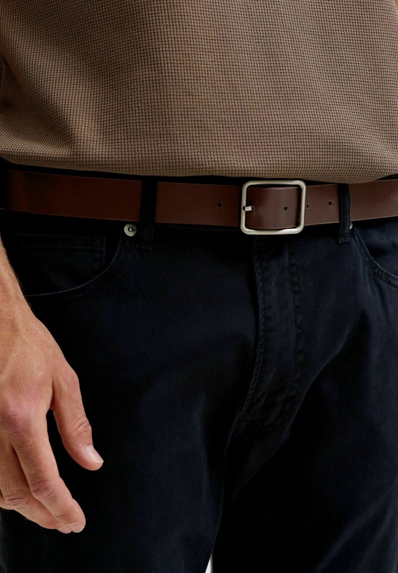 Homme portant une chemise marron texturée rentrée dans un pantalon noir avec une ceinture en cuir marron foncé et une boucle argentée, main détendue le long du corps.