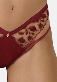 La lingerie bordeaux présente un mesh brodé de fleurs, des parties en tissu lisse et des accents en métal doré qui rehaussent le design.