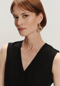 Schwarzes ärmelloses Top mit einem Knopfdetail, kombiniert mit großen goldenen kreisförmigen Ohrringen als kontrastierendes Accessoire. Glatte Textur und klare Linien.