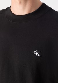 Czarna bawełniana koszulka z okrągłym dekoltem, z małym białym haftem logo "cK" na klatce piersiowej. Teksturowana tkanina, prosty design, bez wzorów.
