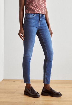 Personne portant un jean skinny bleu, un haut sans manches à motifs, et des mocassins noirs, debout sur un sol en bois devant un mur uni.