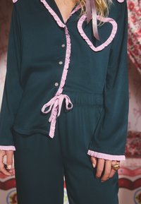 Mörkgrönt pyjamaset med rosa volanger längs kragen, knappslån och manschetter. Har en hjärtformad detalj på bröstet.