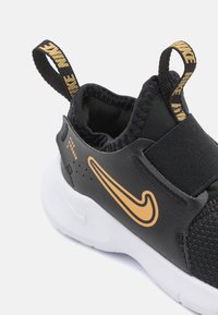 Baskets noires avec un dessus texturé, logo Nike doré et lanière élastique. Semelle blanche avec une finition lisse et une languette pour un enfilage facile.