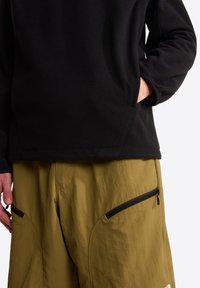 Μαύρο πουλόβερ από φλις με μακριά μανίκια, χαλαρή εφαρμογή και πλαϊνές τσέπες, συνδυασμένο με ελαφριά χακί παντελόνια με τσέπες με φερμουάρ.