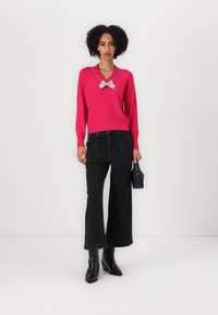 Lys rosa genser med V-hals og blå sløyfeaccent, parret med vide svarte jeans og svarte hæl støvler. Enkel svart håndveske.