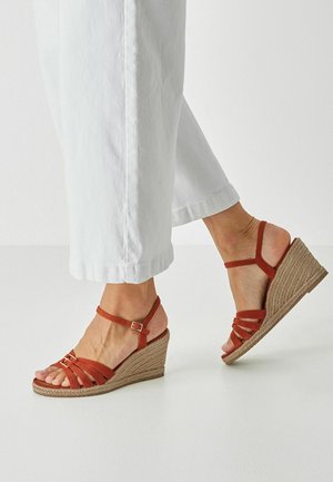 Otevřené klínové sandály rezavé barvy s pásky a proplétanou podešví z juty, s přezkou na suchý zip, na pozadí bílých kalhot.