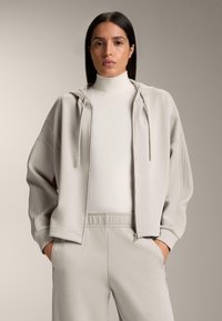 Felpa con cappuccio e zip color grigio chiaro, vestibilità comoda, con dettagli a coste e coulisse. Abbinata a pantaloni grigio chiaro coordinati.