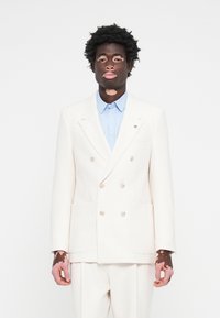 Double-breasted cream suit with large pockets and light blue shirt underneath. Features soft texture and simple buttons.