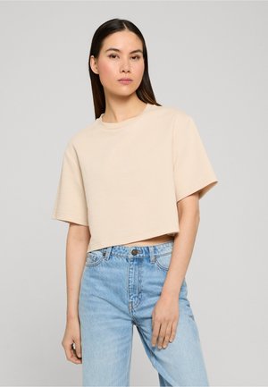 Femme aux longs cheveux foncés portant un haut court beige à manches courtes et un jean taille haute bleu clair, debout devant un fond gris uni.