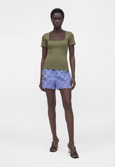 Γυναίκα που φοράει ένα ελαφρώς πράσινο κοντομάνικο ριμπ τοπ, μπλε και άσπρες ριγέ σορτς με κεντήματα με λουλούδια και μαύρα σανδάλια με τακούνι.