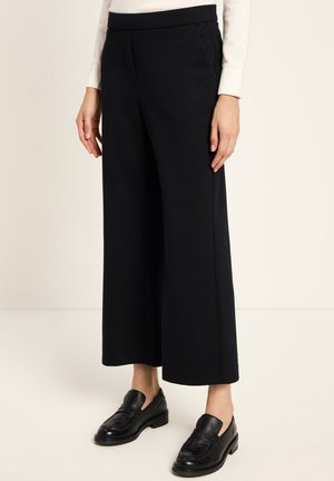 Frau in schwarzen, weit geschnittenen, cropped Hosen, weißem Langarmoberteil und schwarzen Lederloafers, die auf einem hellen Boden steht.