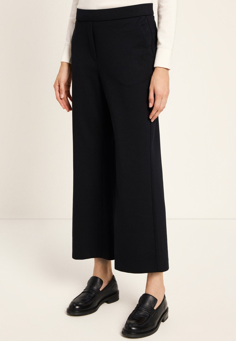 Woman wearing black wide-leg cropped trousers, white long-sleeve top, and black leather loafers standing on a light floor.