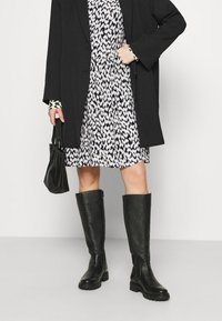 Svarta läderstövlar upp till knäna med strukturerad yta, stabil sula och enkel design, i kombination med en mönstrad klänning och svart kavaj.