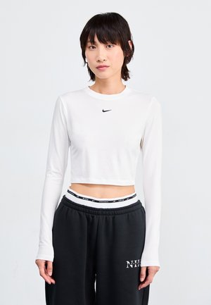 Bijela skraćena duga majica s dugim rukavima i okruglim izrezom, s crnim Nike swoosh logotipom. Nosila se s crnim trenirkama koje imaju brendirani pojas.