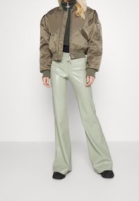 Giacca bomber in satin verde oliva con polsini a coste, abbinata a pantaloni a palazzo lucidi di un verde chiaro. Entrambi i capi hanno dettagli di design minimalisti.
