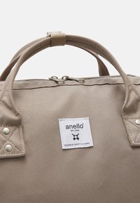 Beige Anello stoffen tas met dubbele handvatten, metalen rits en een wit logo-label met de tekst "anello EST. 2005, ontworpen door Carrot Co. in Japan."