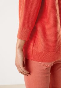 Maglione lavorato a maglia corallo con orlo e maniche a costine, abbinato a jeans corallo rosa. La texture appare morbida e leggermente elastica.