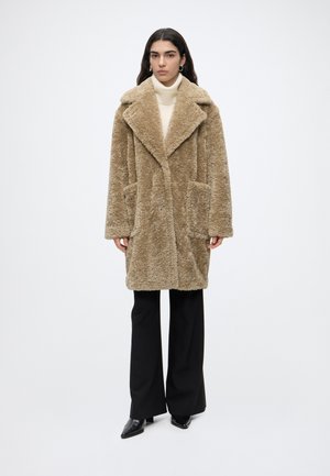 Femme portant un manteau teddy couleur beige sur un col roulé blanc, un pantalon large noir et des chaussures noires pointues, debout devant un fond uni.