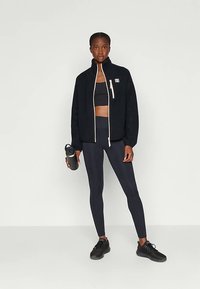 Donna in abbigliamento sportivo nero, con una bottiglia d'acqua nera in mano, indossa una giacca con zip, leggings, top corto e sneakers.