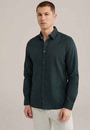 Camisa de botones de manga larga verde oscuro en tela texturizada, con cuello y puños, combinada con pantalones de color claro.