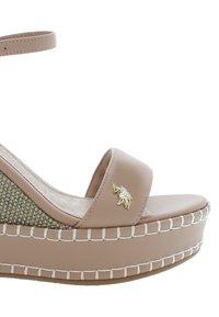 Béžové wedge sandály s širokým páskem, texturovaným tkaným akcentem, zlatým emblémem a bílými stehy kolem podešve. Plochý základ s platformou.