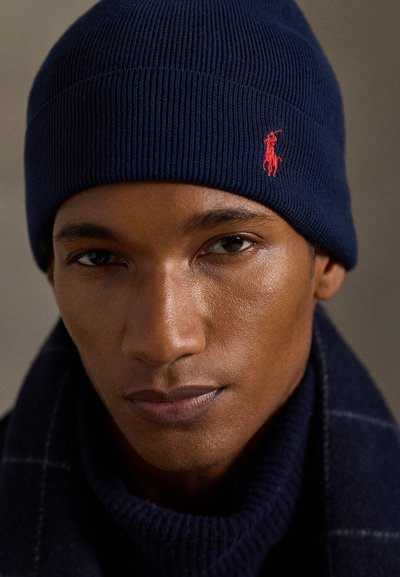 Gorro de malha canelada azul-marinho com um pequeno logotipo vermelho bordado. Tem uma textura suave e um ajuste confortável, concebido para aquecer e proporcionar conforto.
