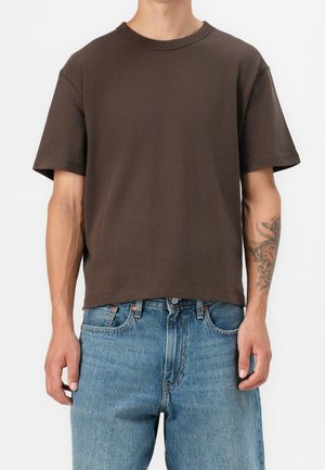 Mand iført en simpel mørkebrun t-shirt med korte ærmer og lyseblå denimjeans, med en blomster-tatovering synlig på venstre underarm.