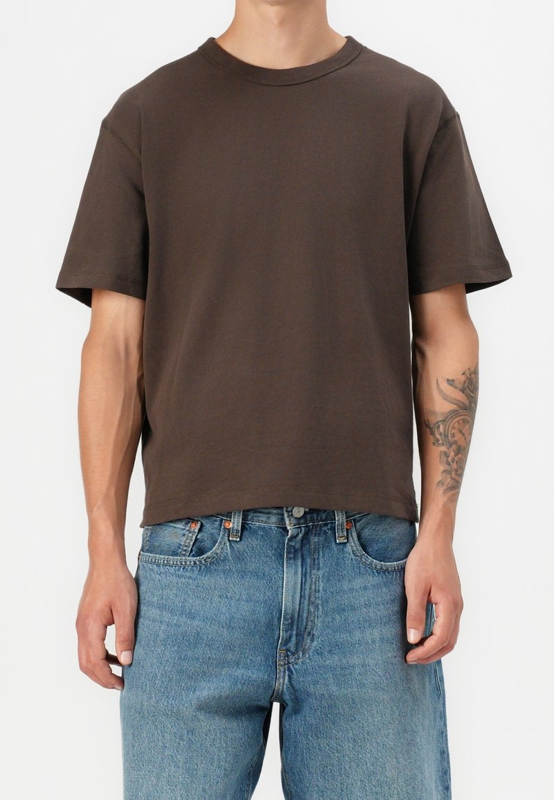 Homme portant un t-shirt uni marron foncé à manches courtes et un jean en denim bleu clair, avec un tatouage floral visible sur l'avant-bras gauche.