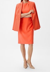Blazer ajusté orange par-dessus une robe moulante assortie. Les deux vêtements ont une texture lisse. Inclut des chaussures à talons hauts beiges.