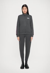 Giacca grigia con zip e colletto alto, abbinata a pantaloni tapered coordinati. Texture liscia, stile aderente e stivali alla caviglia neri.