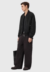 Μαύρο μπουφάν τύπου bomber με φερμουάρ μπροστά και ελαστικά μανίκια, σε συνδυασμό με μαύρο παντελόνι φαρδύ και σκούρα παπούτσια. Κρατάει μια υφασμάτινη μαύρη τσάντα tote.